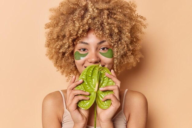 Happy young woman covers mouth with green calla wears hydrogel patches under eyes has curly bushy hair stands bare shoulders isolated over beige background. Beauty wellness and skin care concept