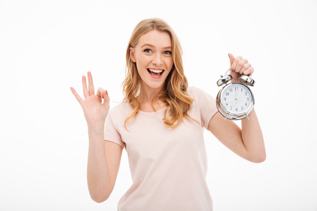 Free photo happy young woman holding alarm clock showing ok gesture.