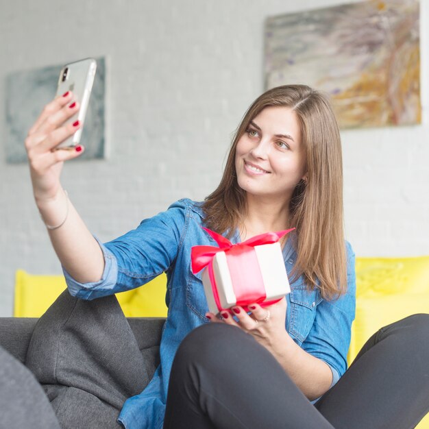 Happy young woman holding birthday present taking selfie with mobile phone