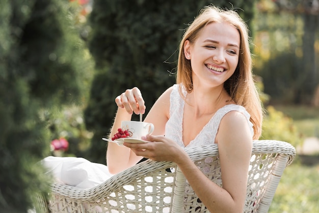 Free photo happy young woman sitting on white chair stirring coffee with spoon