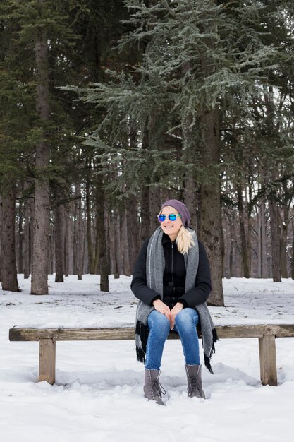 Happy young woman sitting on wooden bench wearing warm clothes in winter