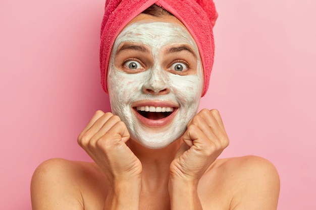 Happy young woman smiles broadly, keeps both hands under chin, cannot believe her eyes, applies clay mask, has joyful face expression