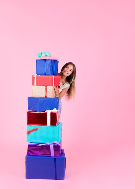 Happy young woman standing behind the stack of different gift boxes on pink backdrop