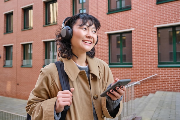 Free photo happy young woman stands on street with backpack and smartphone listens music in headphones waits so