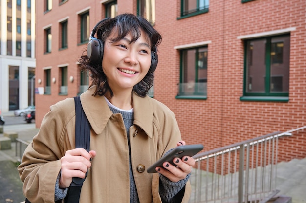 Free photo happy young woman stands on street with backpack and smartphone listens music in headphones waits so