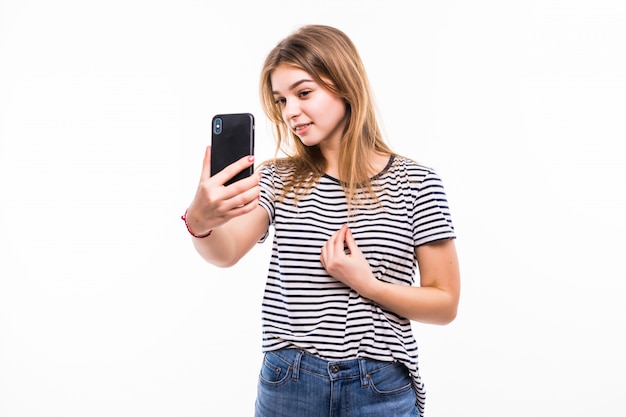 Happy young woman taking pictures of herself at smart phone, over white wall