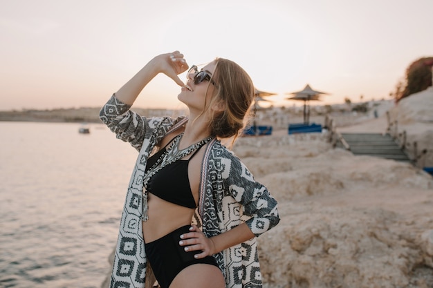 Free photo happy young woman on vacation, shoot on the beach in sunset, posing, biting finger. wearing black swimsuit, sunglasses, necklace, cardigan. enjoying sunset, holiday summertime