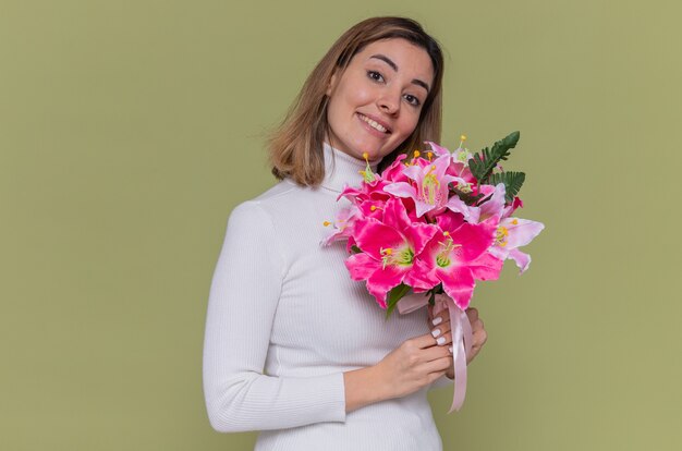 Happy young woman in white turtleneck holding bouquet of flowers looking at front smiling cheerfully celebrating international women's day standing over green wall