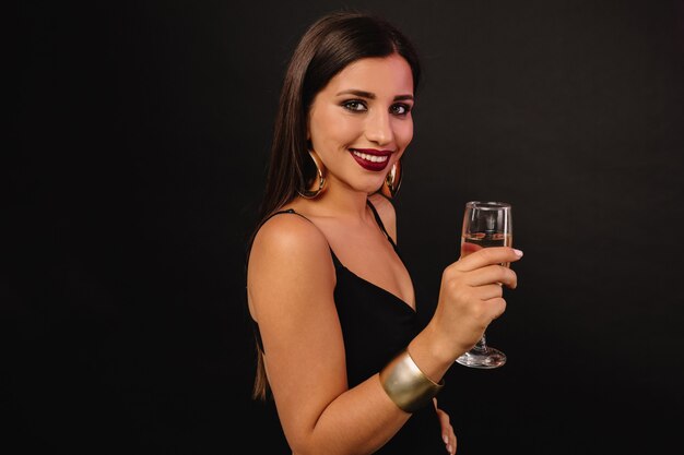 Happy young woman with golden jewerly in black dress drinking champagne