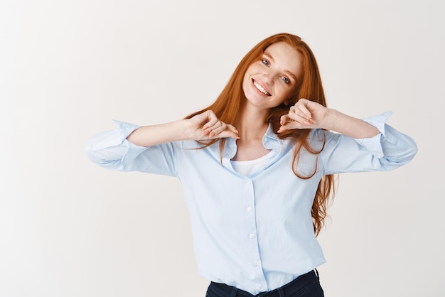 Happy young woman with long red hiar and blue eyes stretching hands smiling cheerfully at camera standing against white background