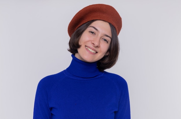 Happy young woman with short hair in blue turtleneck wearing beret looking at front smiling cheerfully standing over white wall