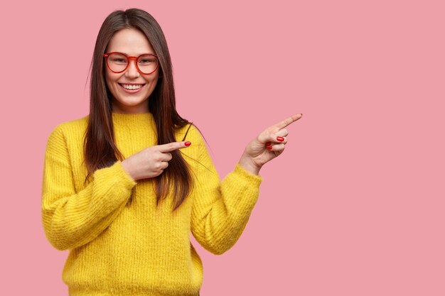 Happy young woman with straight dark hair points aside, advertises new item in clothes shop, dressed in bright yellow sweater