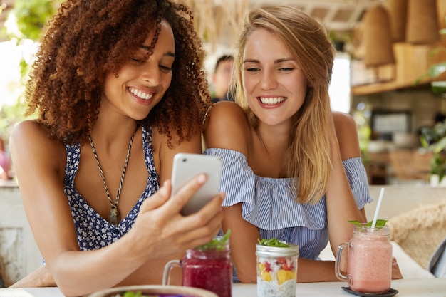 Happy young women watch funny video in internet on smart phone, sit together against cafe interior, eat tasty dessert and fresh cocktails. People, relationship and modern technology concept.