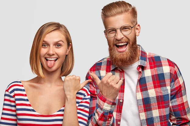 Free photo happy youngsters have fun together, point at each other with joyful expressions. funny cute female shows tongue, overjoyed bearded male hipster in checkered shirt, isolated over white wall