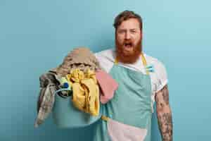 Free photo hard working ginger bearded man does domestic chores, busy with washing, holds basket of laundry, wears apron, clothespins, exclaims loudly, overstained with housekeeping. household concept.