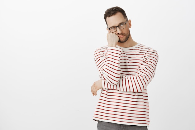 Hate waiting in queues. Portrait of annoyed tired handsome guy with bristle in black eyewear, leaning face on hand and looking indifferent, standing bored over gray wall