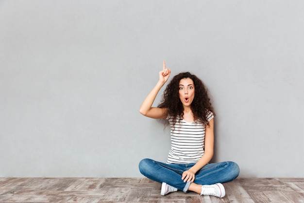 Free photo have idea! cute woman in casual clothes sitting with legs crossed on the floor gesturing index finger up meaning eureka over grey wall