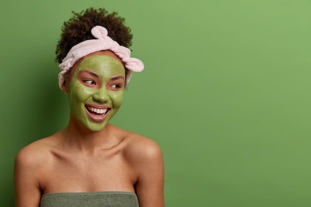 Head shot of smiling carefree woman has bare shoulders, looks away with pleasure, does skin care beauty facial routine, wears green mask on face, headband, isolated over  wall with blank space