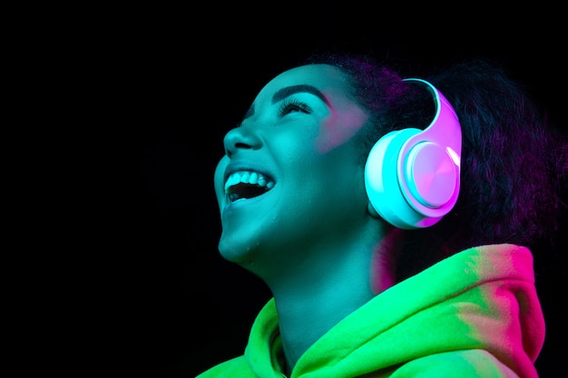 Headphones. African-american woman's portrait isolated on dark studio background in multicolored neon light. Beautiful female model. Concept of human emotions, facial expression, sales, ad, fashion.