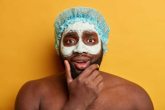 Headshot of Afro man holds chin, applies clay mud mask for having healthy skin, looks with worried expression, wears shower cap