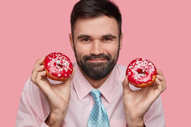 Free photo headshot of attractive bearded male has friendly expression, gentle smile, thick bristle, wears shirt and tie, holds tasty doughnuts in both hands