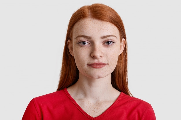 Headshot of attractive red haired European woman with freckled skin, looks seriously at camera, has minimal make up, wears red sweater, isolated on white. Natural beauty concept