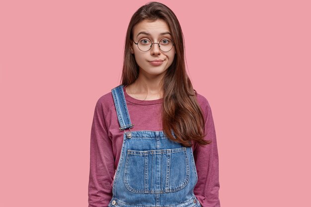 Headshot of beautiful brunette young lady thinks about problem, demonstrates confused emotion, wears denim dungarees, purses lips