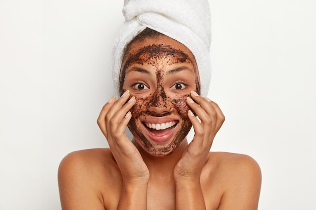 Free photo headshot of beautiful dark skinned woman wears coffee scrub facial mask, keeps hands on cheeks, smiles broadly, wears white towel on head, models indoor against white wall. face treatment