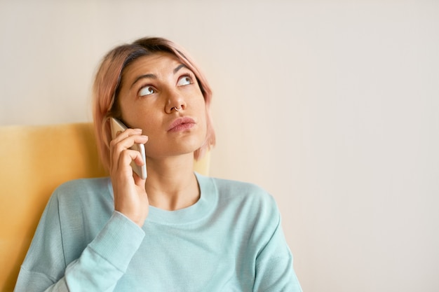 Free photo headshot of beautiful teenage girl with nose piercing looking up with thoughtful facial expression while speaking on cell phone, trying to remember something.