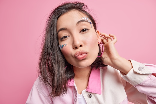 Free photo headshot of beautiful young asian woman keeps lips rounded gazes romantic  flirts with handsome man tilts head has natural beauty wears stylish clothing models against pink wall