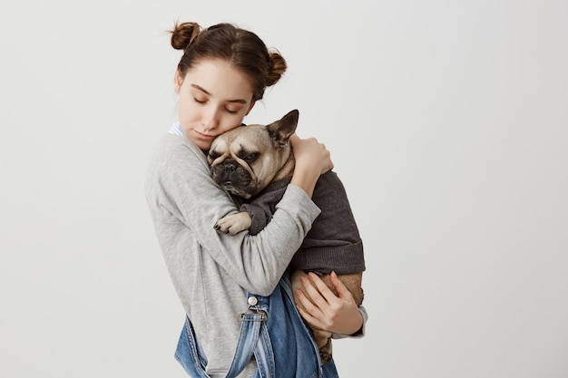 Free Photo headshot of caucasian woman with closed eyes holding her lovely pet like child relaxing together. tender emotions of cute girl cuddling her small dog dressed in sweater. care, love concept