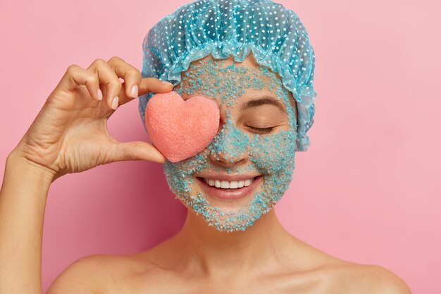 Headshot of cheerful young woman with crystal sea salt scrub, holds pink heart shaped sponge on eye, smiles positively, wears showercap, models against pink wall, peels face from pores