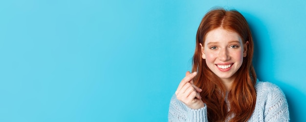 Headshot of cute caucasian woman with red hair and freckles showing heart sign and smiling standing