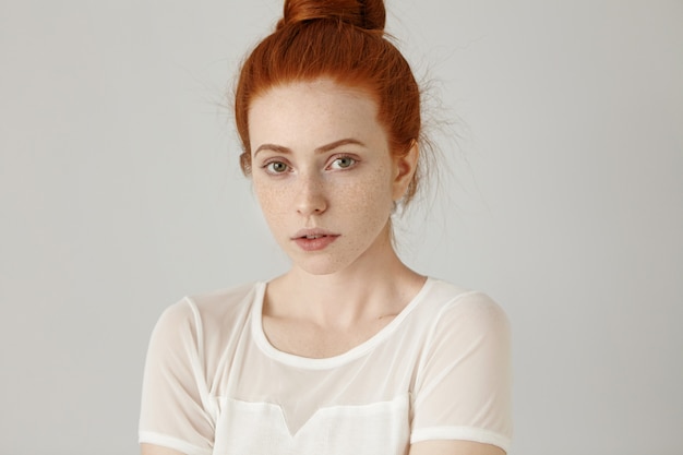 Headshot of cute girl with pleasant features posing at grey wall. Attractive young Caucasian woman with ginger hair in bun and freckles looking and smiling with her lips slightly parted