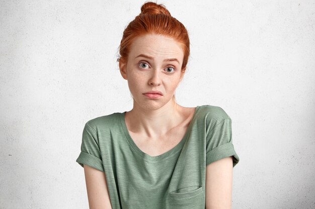 Headshot of discontent uncertain doubtful female with red hair tied in bun, hesitates about something, being displeased with everything poses against white