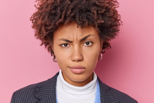 Free photo headshot of displeased young curly haired woman frowns face raises eyebrows dressed in elegant clothes listens something attentively isolated over pink background. negative human emotions concept