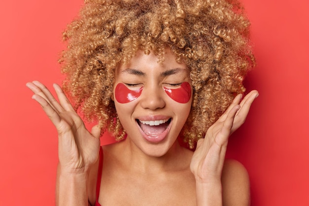 Headshot of emotional curly haired blonde young woman raises hands near head exclaims loudly keeps eyes closed applies hydrogel patches under eyes undergoes beauty treatments. Wellness concept