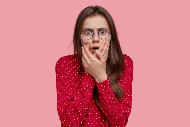 Headshot of frightened lady looks with terrible expression, keeps hands near mouth, hears bad news, has dark hair