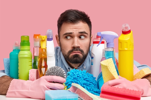 Headshot of handsome bearded man focused aside, has puzzled look, surrounded with cleaning detergents