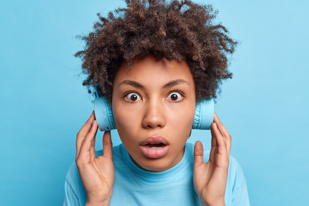 Headshot of impressed stunned curly haired woman has eyes popped out keeps breath listens music in wireless headphones shocked to hear very loud sound unexpectedly isolated on blue  wall