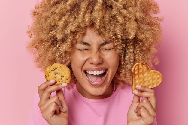 Free photo headshot of joyful european woman with curly hair holds delicious waffle and cookie enjoys eating homemade desserts exclaims from happiness wears casual t shirt isolated over pink background