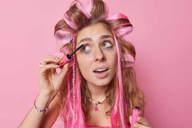 Free Photo headshot of long haired young european woman applies hair rollers and mascara looks away undergoes beauty treatments prepares for first date wants to look beautiful isolated over pink background