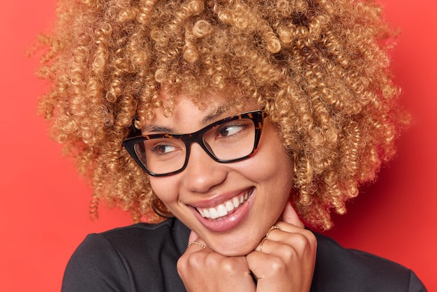 Free photo headshot of lovely cheerful woman smiles pleasantly keeps hands under chin has charming glance away wears optical spectacles poses against vivid red background positive human face expressions