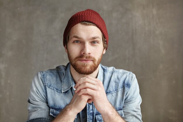 Headshot of modern good-looking young European hipster wearing stylish hat and blue jeans shirt holding hands clasped in front of him, having thoughtful and dreamy look, sitting at concrete wall