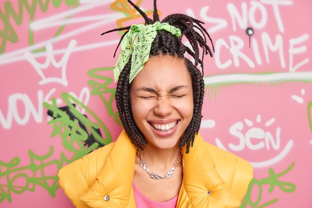 Headshot of overjoyed woman closes eyes smiles toothily has fun in urban place wears stylish clothes poses against street graffiti wall expresses positive emotions