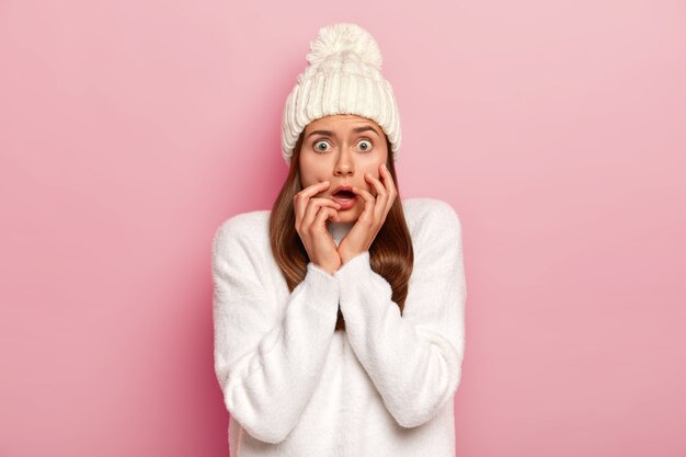 Headshot of perplexed astonished European woman popping eyes and has scared look, wears white winter outfit, has desperate frightened expression, thinks anxiously