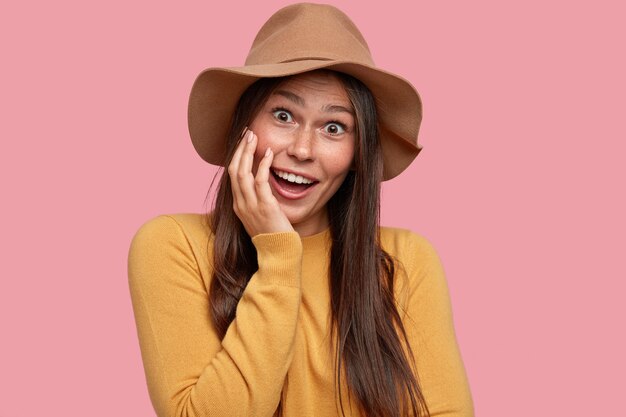 Headshot of positive freckled young woman with satisfied expression, keeps hand on cheek