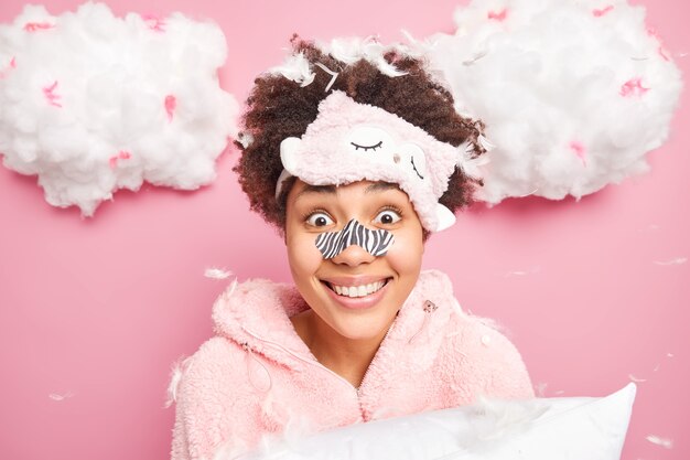 Headshot of positive woman with curly hair wears blindfold nose patch slumber suit hears amazing news holds pillow isolated over pink wall