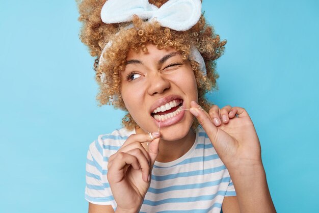 Headshot of pretty woman cleans teeth with dental floss undergoes daily hygiene routines winks eye removes food after eating wears casual striped t shirt isolated over blue wall Dental care