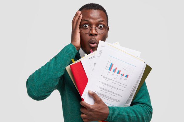 Headshot of puzzled African American man touches head, stares with bugged eyes, opens mouth from amazement, holds paper documents